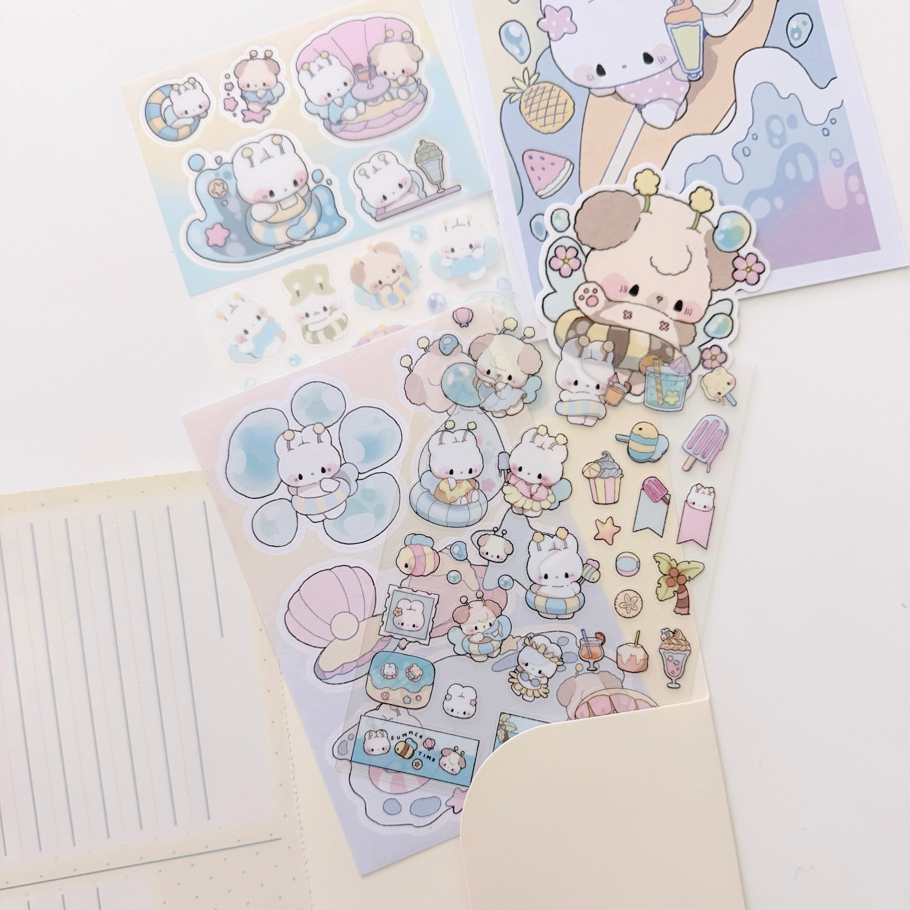 A set of cute pastel sticker sheets featuring a small white cat character in various playful outfits and accessories, arranged on a white desk next to a lined notepad and an envelope.